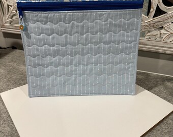 May include: A square, quilted fabric storage container. The top section features a blue and white patterned fabric, while the main body is light blue with vertical white stitching. A blue zipper and a small charm are visible.