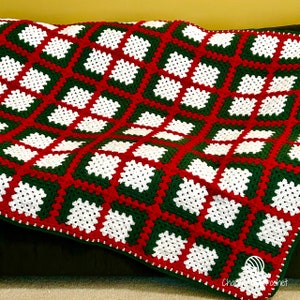 May include: A handmade crochet blanket with a repeating pattern of white squares framed by green and red. The blanket is made with a textured stitch and has a green border. The blanket is folded over a dark surface.