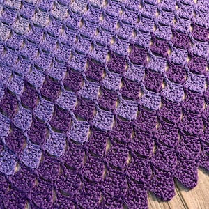 May include: A purple and lavender crochet blanket with a scalloped edge. The blanket is made with a repeating pattern of small, overlapping triangles.