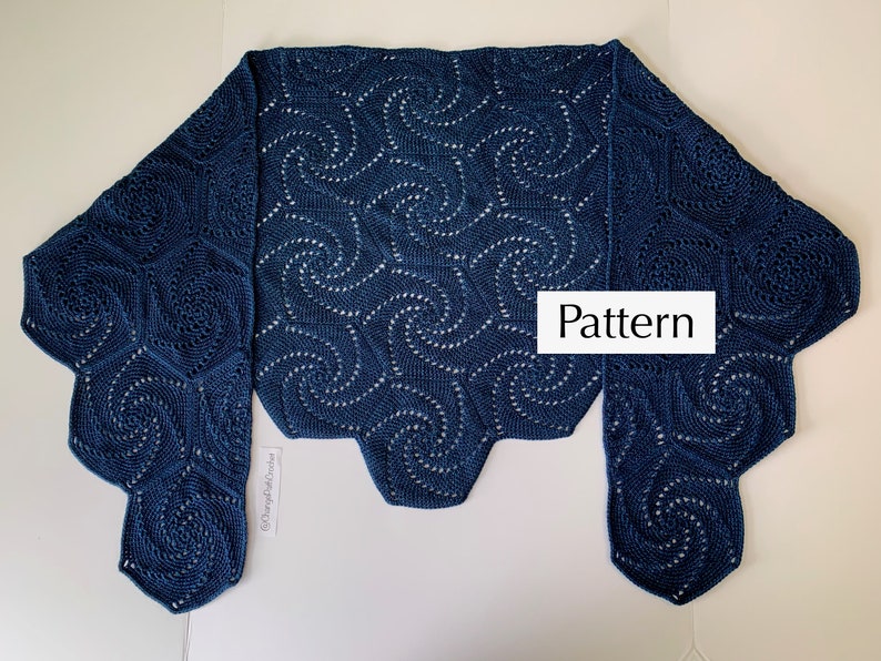 A crochet shawl lies flat on a surface with the two ends folded over. The shawl is made of hexagons with a swirl pattern.