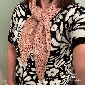 May include: A crocheted scarf in a light pink colour with a lacy pattern. The scarf is tied around the neck of a person wearing a black and white floral shirt.