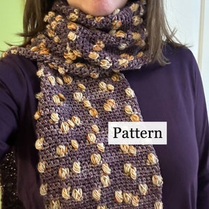 May include: A purple and beige crocheted scarf with a textured pattern. The scarf is folded in half and draped over a person's neck. The text "Pattern" is visible on the scarf.