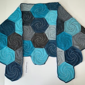 A crochet shawl lies flat on a surface with the two ends folded over. The shawl is made of hexagons with a swirl pattern. The hexagons are all different shades of blue.