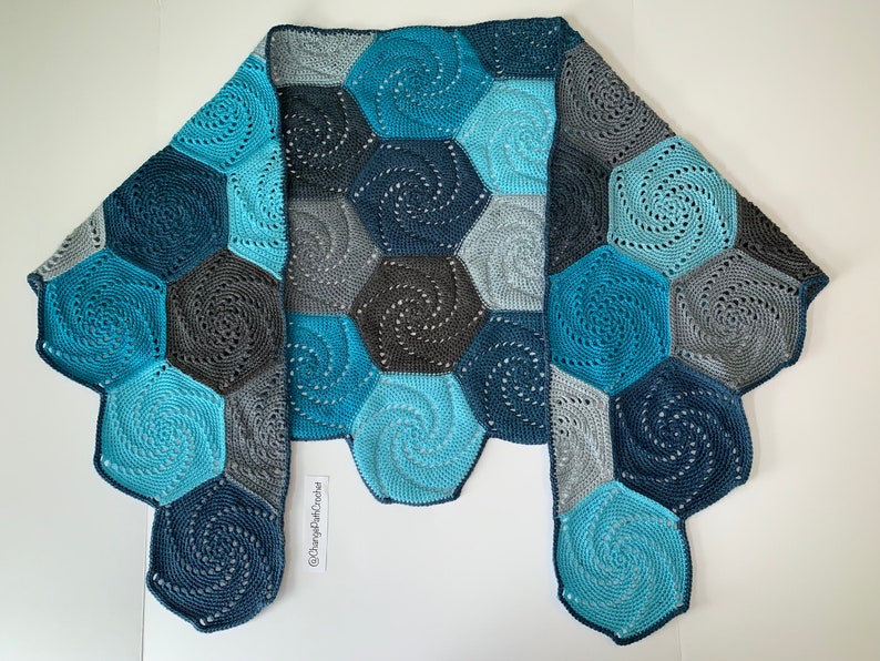 A crochet shawl lies flat on a surface with the two ends folded over. The shawl is made of hexagons with a swirl pattern. The hexagons are all different shades of blue.