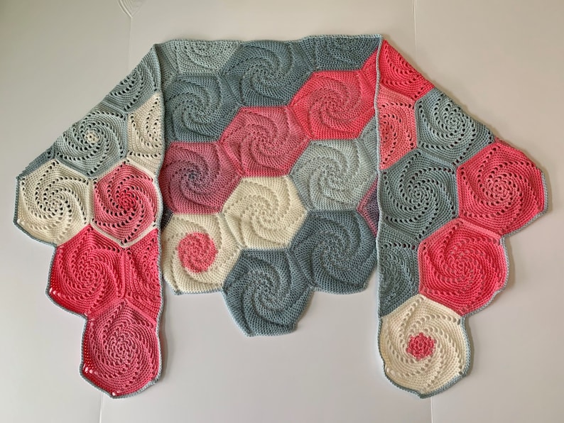 A crochet shawl lies flat on a surface with the two ends folded over. The shawl is made of hexagons with a swirl pattern. The hexagons are all different shades of pink and gray.