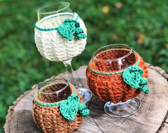 May include: Three crocheted pumpkin wine glass cozies in white, orange, and brown with green leaf accents. The cozies are sitting on a tree stump.