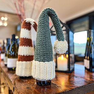 Op de afbeelding: Twee wijnflessen met gebreide flessenhoezen. De flessenhoezen zijn groen-wit gestreept en groen met een witte pompon.