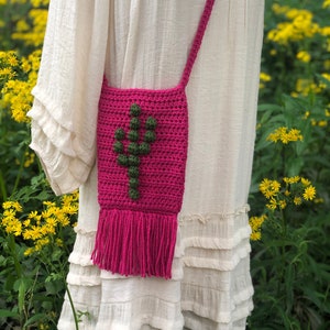 Puede incluir: Un bolso de hombro de crochet rosa con un diseño de cactus verde y flecos. El bolso lo lleva una persona que lleva una blusa de crochet blanca.