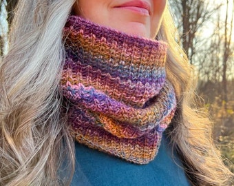 May include: A woman wearing a teal blue shirt and a multi-colored knitted neck warmer. The neck warmer has shades of purple, pink, brown, and grey. The woman is looking up and smiling. The background is a blurry image of trees and a sunset.