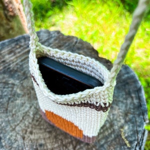 May include: A crocheted phone pouch with a brown, white, and orange striped design. The pouch is hanging from a rope strap and has a black phone inside.