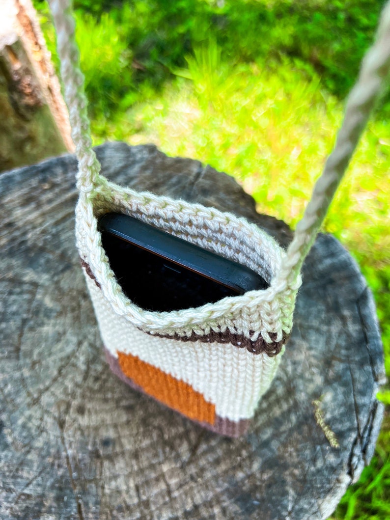 May include: A crocheted phone pouch with a brown, white, and orange striped design. The pouch is hanging from a rope strap and has a black phone inside.