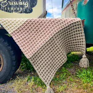 Op de afbeelding: Een driehoekig gehaakt deken met een wafelpatroon in drie kleuren: beige, lichtroze en cr&egrave;me. Het deken heeft twee kwastjes aan de onderste hoeken.