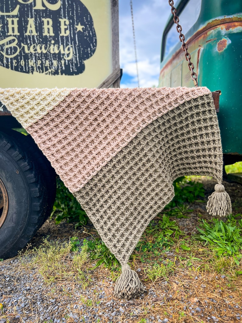 Op de afbeelding: Een driehoekig gehaakt deken met een wafelpatroon in drie kleuren: beige, lichtroze en cr&egrave;me. Het deken heeft twee kwastjes aan de onderste hoeken.