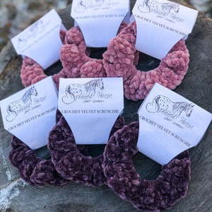 May include: Five crochet velvet scrunchies in two colors, pink and purple, with a white label that says "Spotted Horse Craft Company" and "Crochet Velvet Scrunchie".
