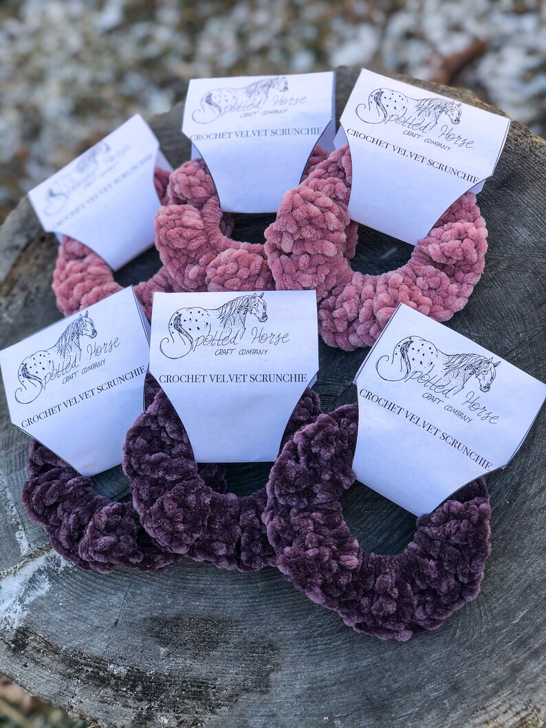 May include: Five crochet velvet scrunchies in two colors, pink and purple, with a white label that says "Spotted Horse Craft Company" and "Crochet Velvet Scrunchie".