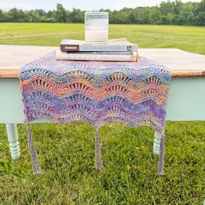 May include: A rectangular crochet table runner featuring a multicolored wavy pattern in shades of purple, blue, orange, and yellow. The runner has three decorative tassels hanging from the front edge and is topped with two books and a glass candle.