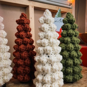 May include: Three crocheted Christmas trees in different colors: white, brown, and green. The trees are made of small, round balls and are arranged in a row on a wooden surface.