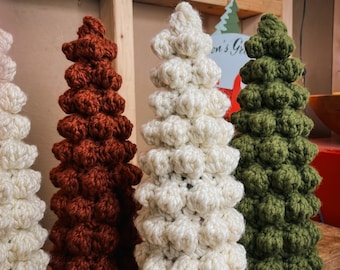 Op de afbeelding: Drie gehaakte kerstboompjes in verschillende kleuren: wit, bruin en groen. De boompjes zijn gemaakt van kleine, ronde balletjes en zijn in een rij op een houten oppervlak gerangschikt.