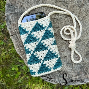Puede incluir: Una funda de tel&eacute;fono de ganchillo con un patr&oacute;n geom&eacute;trico de color verde azulado y blanco. La funda tiene una larga correa blanca para llevarla al hombro.