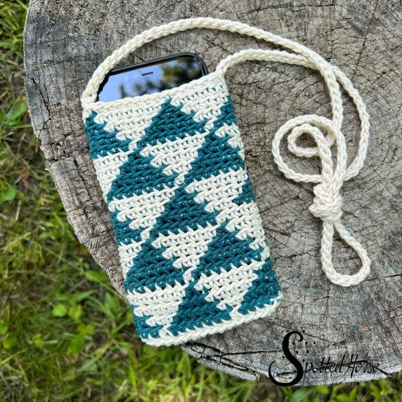 Puede incluir: Una funda de tel&eacute;fono de ganchillo con un patr&oacute;n geom&eacute;trico de color verde azulado y blanco. La funda tiene una larga correa blanca para llevarla al hombro.