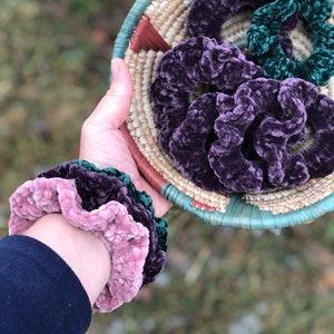 May include: A collection of purple and green velvet scrunchies in a woven basket. The scrunchies are soft and fluffy, and they would make a great gift for anyone who loves hair accessories.