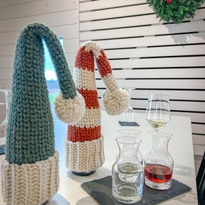 Puede incluir: Dos botellas de vino con cubiertas de botellas de punto en forma de gorros de elfo. Los gorros son verde y blanco y rojo y blanco. Las botellas est&aacute;n sobre una mesa blanca con dos peque&ntilde;as jarras de vidrio llenas de vino tinto y blanco.