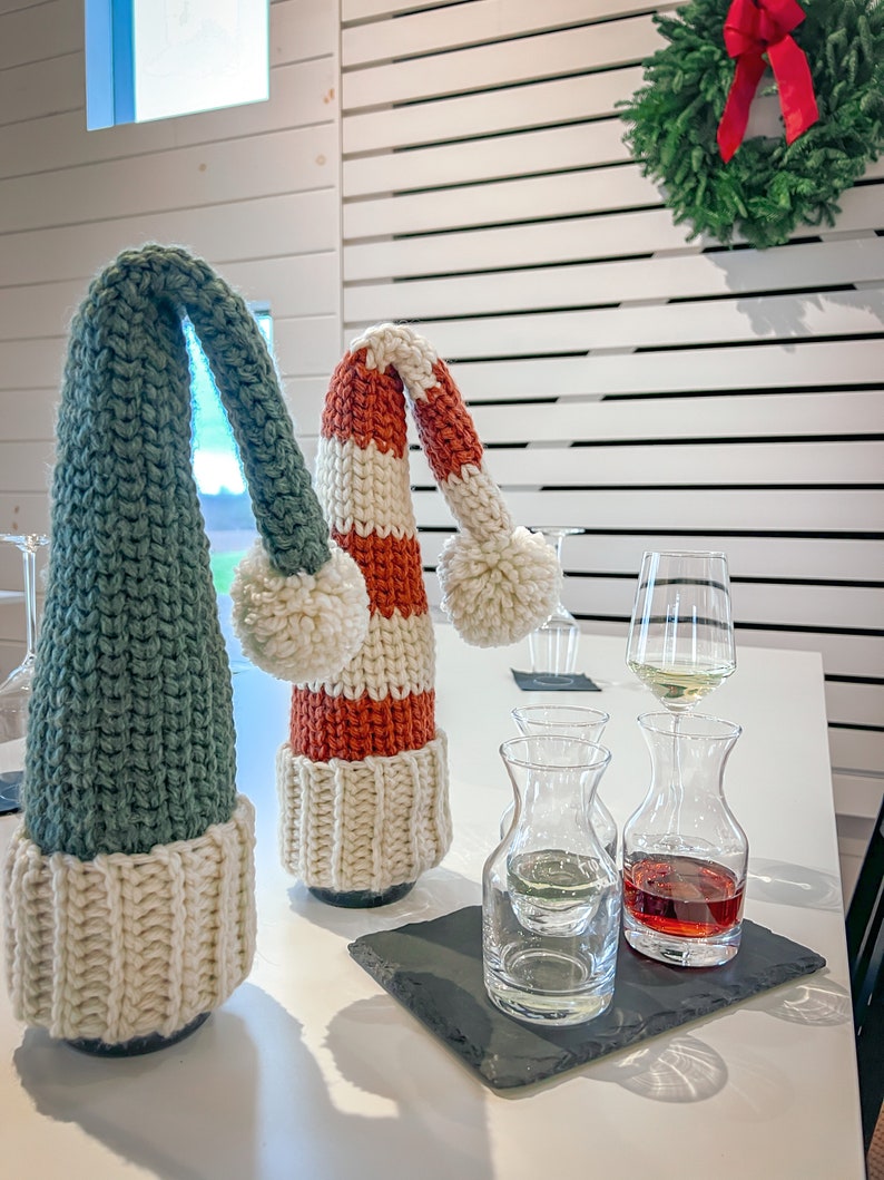 Puede incluir: Dos botellas de vino con cubiertas de botellas de punto en forma de gorros de elfo. Los gorros son verde y blanco y rojo y blanco. Las botellas est&aacute;n sobre una mesa blanca con dos peque&ntilde;as jarras de vidrio llenas de vino tinto y blanco.