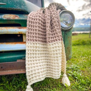 Op de afbeelding: Een gehaakt deken met een wafelpatroon in beige, cr&egrave;me en bruin. Het deken is over de voorkant van een vintage groene vrachtwagen gelegd.