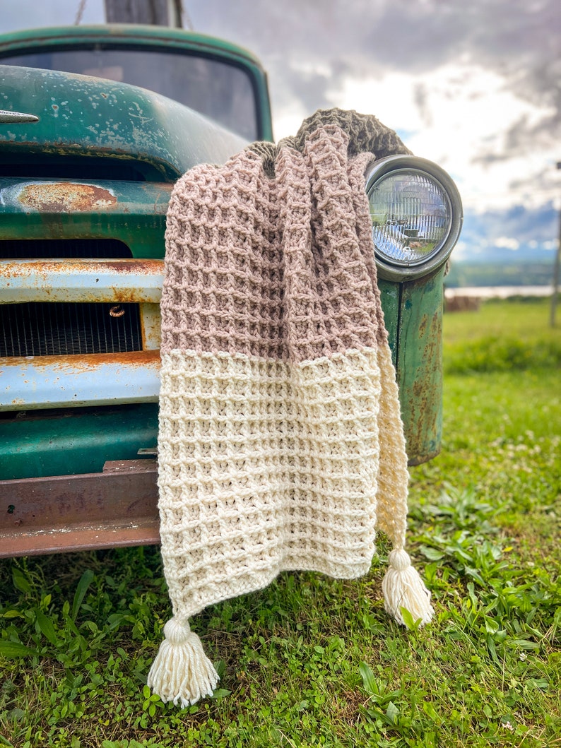 Op de afbeelding: Een gehaakt deken met een wafelpatroon in beige, cr&egrave;me en bruin. Het deken is over de voorkant van een vintage groene vrachtwagen gelegd.