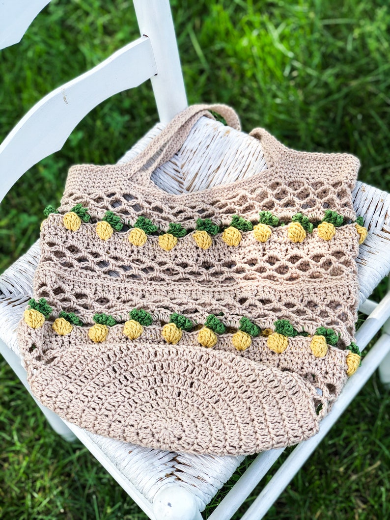 Puede incluir: Una bolsa de mano de crochet con base marr&oacute;n y detalles de pi&ntilde;a amarillos y verdes. La bolsa est&aacute; sentada en una silla blanca.