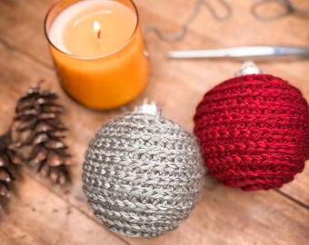 May include: A lit orange candle in a glass jar, two crocheted Christmas ornaments, one silver and one red, and a pine cone rest on a wooden surface. The ornaments are round and have silver metal caps.