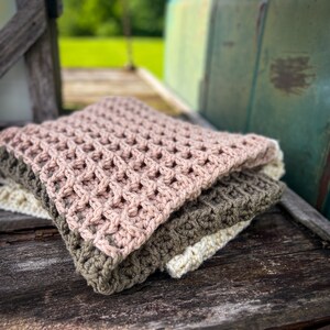 Op de afbeelding: Een stapel van drie gehaakte dekens in roze, groen en wit. De dekens zijn gevouwen en geplaatst op een houten oppervlak.