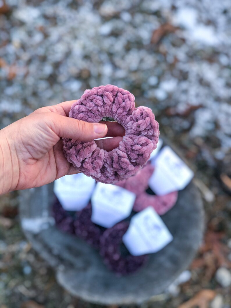 May include: A close-up of a soft, pink, fuzzy hair scrunchie. The scrunchie is made of a thick, textured fabric and is held in a hand.