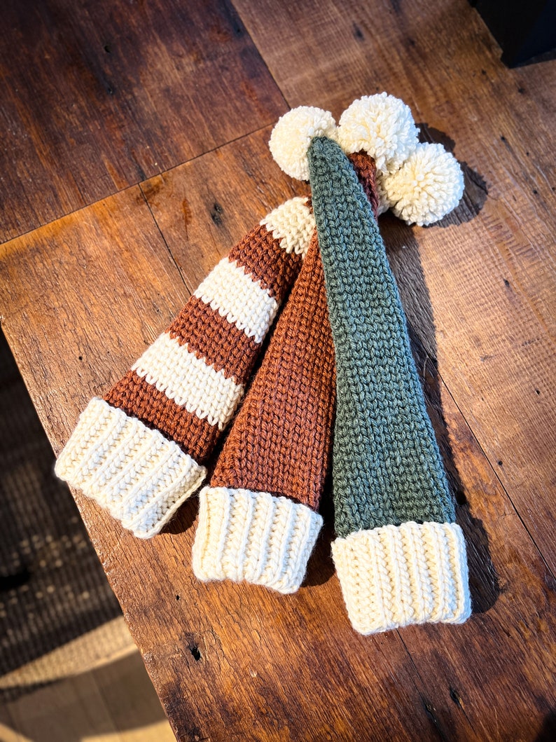 Puede incluir: Tres gorros de punto con pompones. Un gorro es de rayas marrones y blancas, otro es marr&oacute;n y el &uacute;ltimo es verde.