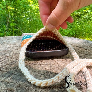 May include: A crocheted phone case with a teal, orange and white striped design. The case is open to reveal a black phone inside. The case has a white crocheted strap.
