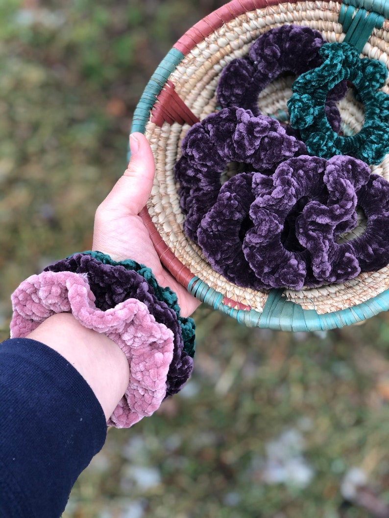 May include: A collection of purple and green velvet scrunchies in a woven basket. The scrunchies are soft and fluffy, and they would make a great gift for anyone who loves hair accessories.