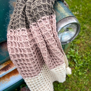 Op de afbeelding: Een gehaakt deken in drie kleuren: bruin, roze en wit. Het deken is gedrapeerd over de voorkant van een vintage groene vrachtwagen.
