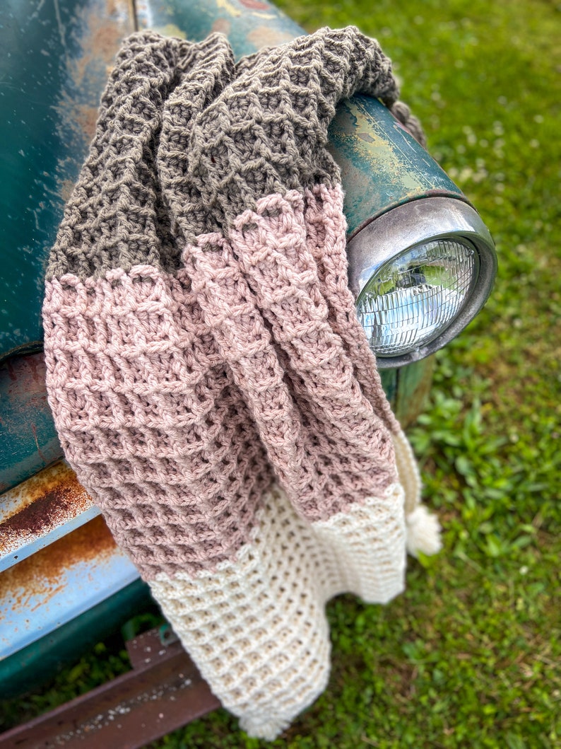Op de afbeelding: Een gehaakt deken in drie kleuren: bruin, roze en wit. Het deken is gedrapeerd over de voorkant van een vintage groene vrachtwagen.