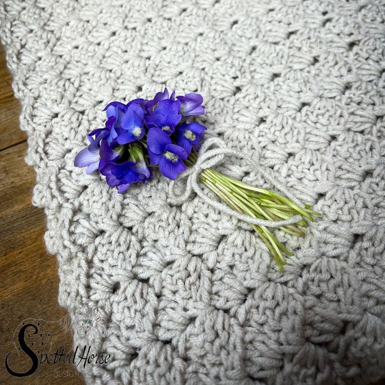 Puede incluir: Un ramo de flores moradas y azules atadas con una cinta blanca, descansando sobre una manta de ganchillo de fondo beige.