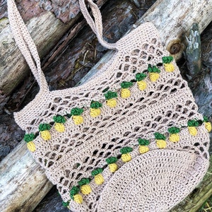 Puede incluir: Una bolsa de mano de crochet con un dise&ntilde;o de pi&ntilde;a. La bolsa es beige con detalles verdes y amarillos. La bolsa est&aacute; sobre una superficie de madera.