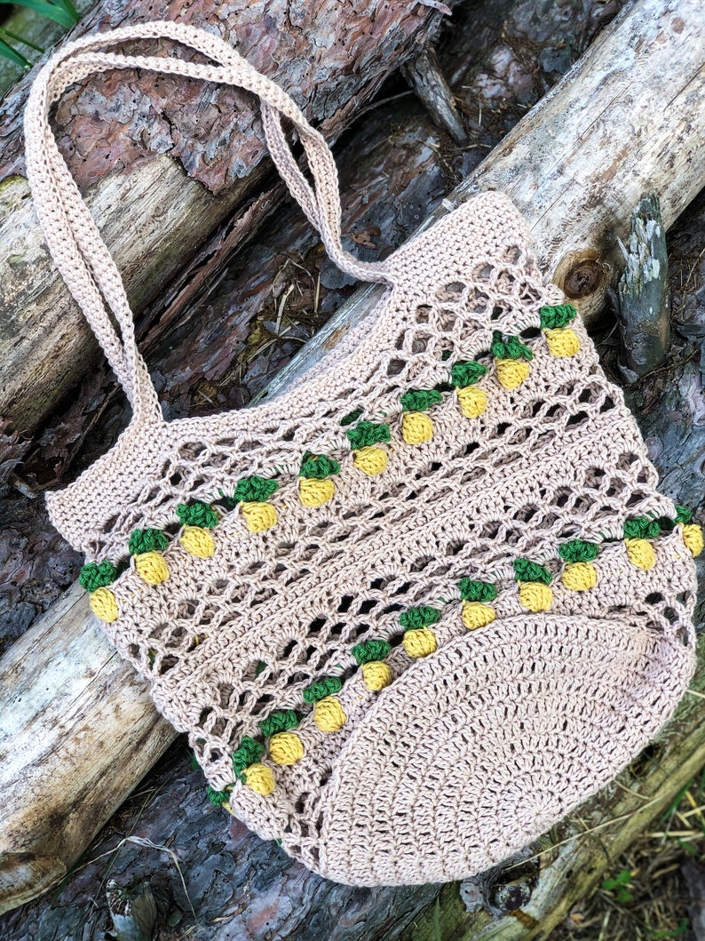 Puede incluir: Una bolsa de mano de crochet con un dise&ntilde;o de pi&ntilde;a. La bolsa es beige con detalles verdes y amarillos. La bolsa est&aacute; sobre una superficie de madera.