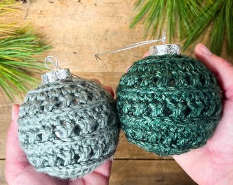 May include: Two crocheted Christmas baubles in shades of green. The baubles are made with a textured stitch and have a silver hanger.