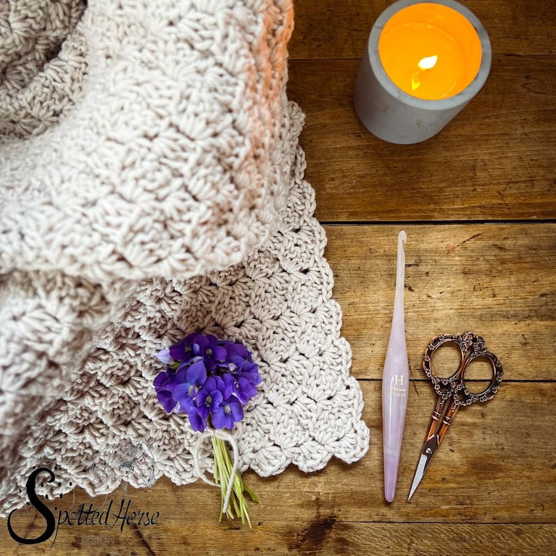 Puede incluir: Una manta de ganchillo de color crema con un borde festoneado. Un ramo de flores moradas descansa sobre la manta. Un gancho de ganchillo rosa y un par de tijeras doradas est&aacute;n sobre una superficie de madera. El texto "Spotted Horse" es visible en la esquina inferior izquierda de la imagen.