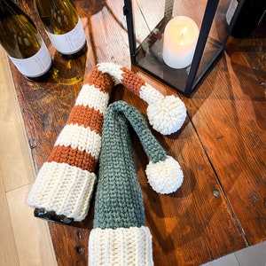 Puede incluir: Dos cubiertas de botella de ganchillo, una con rayas marrones y blancas y otra con rayas verdes y blancas, con pompones blancos en la parte superior. Las cubiertas de botella est&aacute;n sobre una mesa de madera con botellas de vino en el fondo.