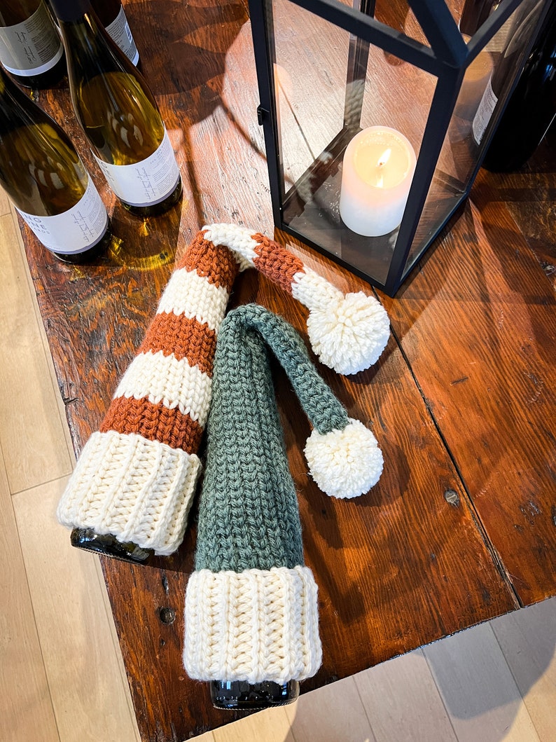 Puede incluir: Dos cubiertas de botella de ganchillo, una con rayas marrones y blancas y otra con rayas verdes y blancas, con pompones blancos en la parte superior. Las cubiertas de botella est&aacute;n sobre una mesa de madera con botellas de vino en el fondo.