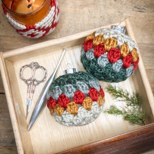 May include: Two crocheted Christmas ornaments in a wooden drawer. The ornaments are green, gray, red, and gold. A crochet hook and a pair of scissors are also in the drawer.
