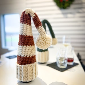 Puede incluir: Un tap&oacute;n de botella de punto de crochet a rayas blancas y marrones con un pomp&oacute;n blanco en la parte superior. El tap&oacute;n de botella est&aacute; sobre una mesa blanca.