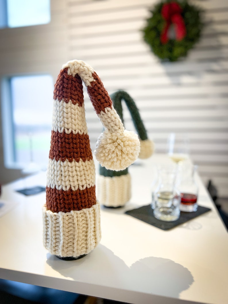 Puede incluir: Un tap&oacute;n de botella de punto de crochet a rayas blancas y marrones con un pomp&oacute;n blanco en la parte superior. El tap&oacute;n de botella est&aacute; sobre una mesa blanca.