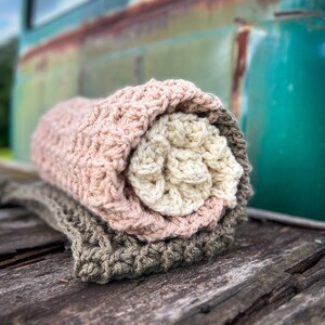 Op de afbeelding: Een stapel van drie gehaakte dekens in tinten roze, cr&egrave;me en bruin. De dekens zijn opgerold en geplaatst op een verweerd houten oppervlak.