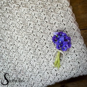 Puede incluir: Primer plano de una manta de ganchillo de color beige claro con un peque&ntilde;o ramo de flores moradas en la parte superior. La manta tiene un patr&oacute;n texturizado en forma de diamante.